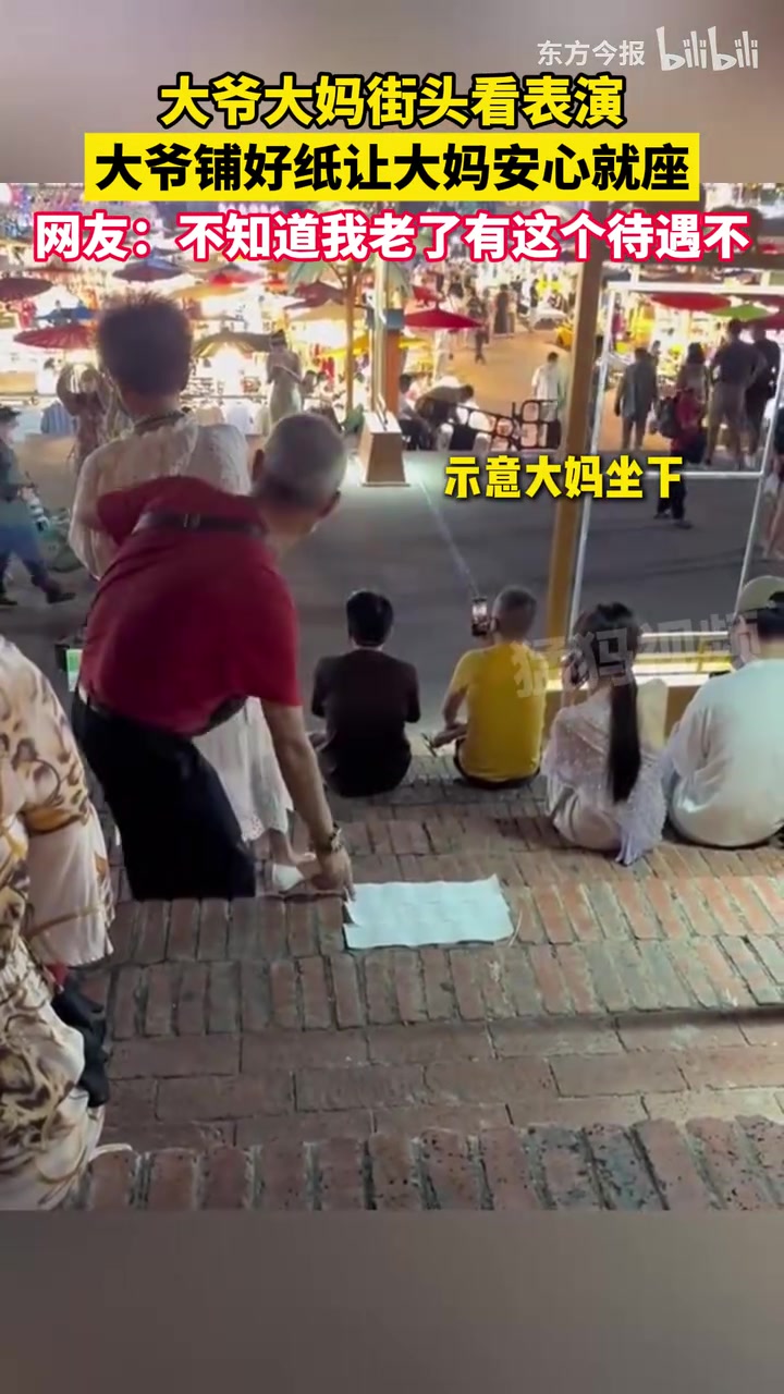 大爷大妈街头看表演,大爷铺好纸让大妈安心就座
