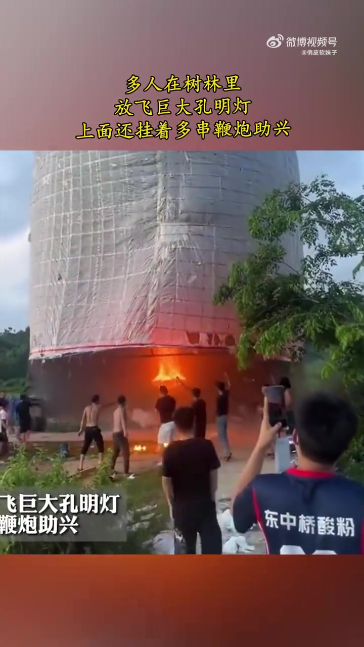 多人在树林里放飞巨大孔明灯，上面还挂着多串鞭炮助兴